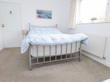 A bedroom with a metal framed bed with blue patterned bedspread a bedside lamp a window with curtains and a radiator at 3 Glyn Y Mor in Llanbedrog