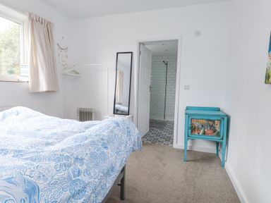 A bedroom with a bed covered in a blue and white patterned duvet a standing mirror a white clothes rack with hangers a small turquoise cabinet and a door leading to a tiled bathroom at 3 Glyn Y Mor in Llanbedrog