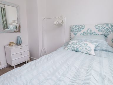 A bedroom with a bed with patterned headboard and pillows a white chest of drawers and a full-length mirror at 3 Glyn Y Mor in Llanbedrog