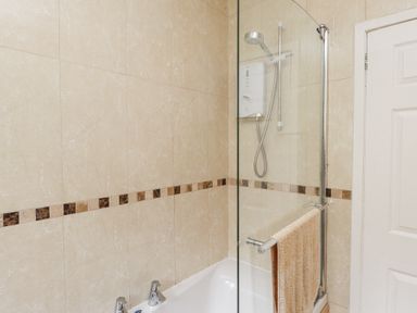 A bathroom with a bathtub shower with glass screen beige tiled walls and a towel on the rail at 3 Glyn Y Mor in Llanbedrog