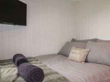 A bedroom with a bed with grey bedding and a pillow that says our place with a wall mounted television at The Highwood Hut near Liskeard