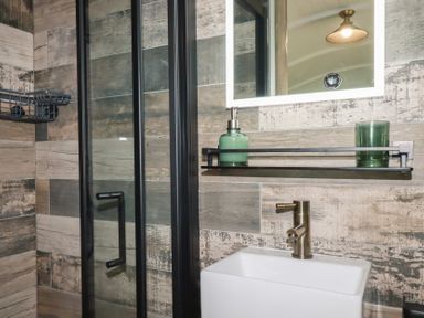 A bathroom with a sink, brass faucet, glass shower enclosure, mirror, and shelf with green soap dispenser and green cup at The Highwood Hut near Liskeard