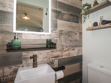 A bathroom with a white sink bronze faucet an illuminated mirror a wooden shelf with green bottles and decorative items and a toilet at The Highwood Hut near Liskeard