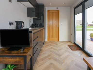 A kitchen area with black cabinets a wooden TV stand with a TV kettle and toaster wooden door and glass sliding doors showing a garden outside at Caban in Dothan near Gwalchmai