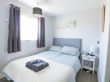 A bedroom with a bed with striped bedding a nightstand with a lamp and clock and a window with curtains at Chalet 38 in Hemsby