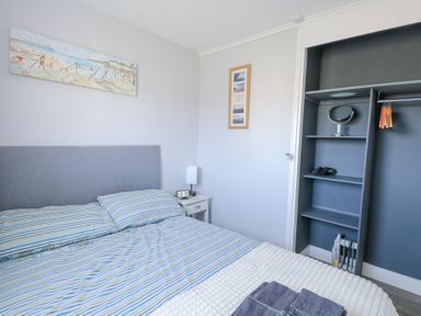 A bedroom with a bed with striped bedding bedside table lamp and clock open closet with hangers and shelves at Chalet 38 in Hemsby