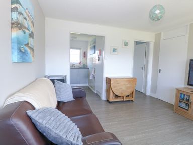 A living room with a brown leather sofa and gray cushions a wooden drop leaf table and a doorway leading to a kitchen at Chalet 39 in Hemsby