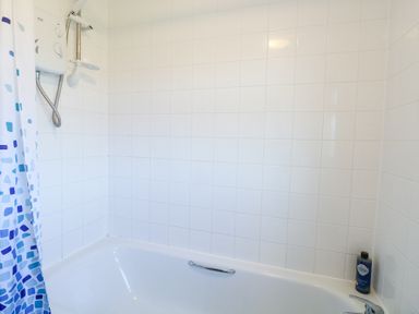 A bathroom with a white bathtub white tiled walls a shower unit blue patterned shower curtain and a bottle on the tub edge at Chalet 39 in Hemsby