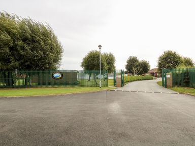 An entrance with a green metal fence and gate opening to a curved driveway with trees and buildings in the background at Chalet 39 in Hemsby
