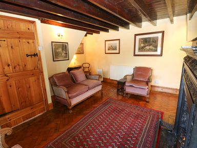 A sitting room with wooden beams a wooden door framed pictures a red patterned rug and upholstered chairs at Pagecroft Cottage in Haydon Bridge
