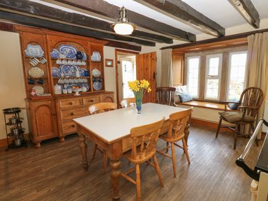 A dining room with a wooden table and chairs a cupboard with blue and white dishes a window seat with cushions and wooden beams on the ceiling at Pagecroft Cottage in Haydon Bridge
