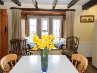 A dining area with a white table and a vase of yellow flowers in front of a window seat with cushions at Pagecroft Cottage in Haydon Bridge