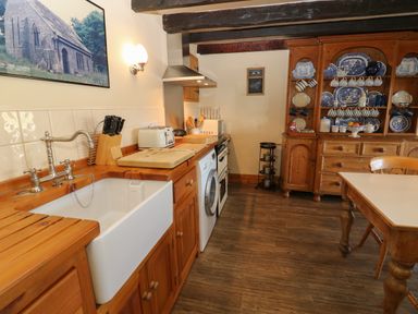 A kitchen with wooden cabinets a white sink a washing machine a stove a wooden dresser with blue and white dishes and a wooden table with a chair at Pagecroft Cottage in Haydon Bridge