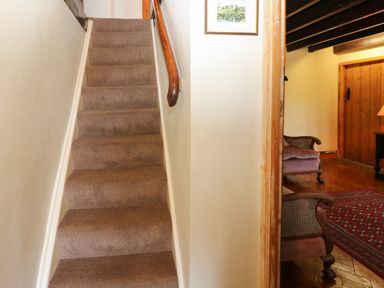 A carpeted staircase with a wooden handrail next to a room with armchairs a rug and wooden door at Pagecroft Cottage in Haydon Bridge