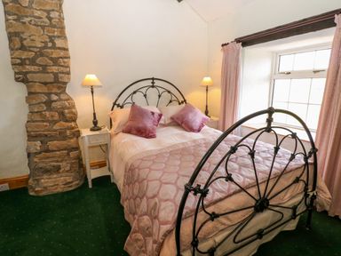 A bedroom with a metal framed bed two bedside tables with lamps a stone feature wall and a window with curtains at Pagecroft Cottage in Haydon Bridge