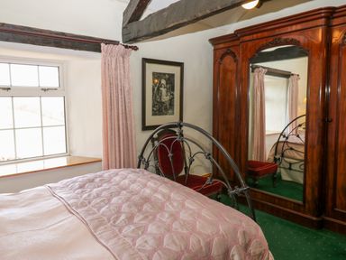 A bedroom with a metal bed frame pink bedding a wooden wardrobe with mirror a red chair and window with pink curtains at Pagecroft Cottage in Haydon Bridge