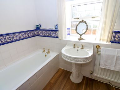 A bathroom with a bathtub a pedestal sink a window with curtains and decorative blue tiles at Pagecroft Cottage in Haydon Bridge