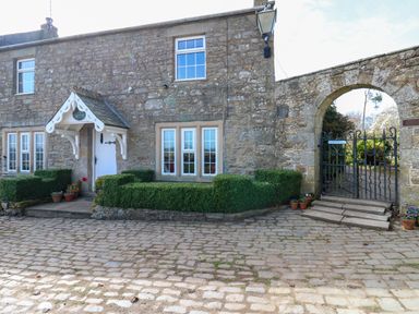 A stone cottage with windows and a white door next to a gated stone archway and cobblestone driveway at Pagecroft Cottage in Haydon Bridge