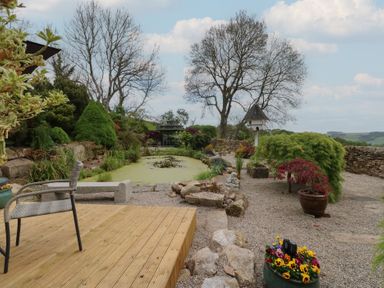 A garden with a pond surrounded by plants and trees and a patio with chairs at Pagecroft Cottage in Haydon Bridge
