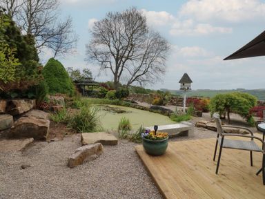 A garden with a pond a stone bench plants a birdhouse and outdoor chairs at Pagecroft Cottage in Haydon Bridge