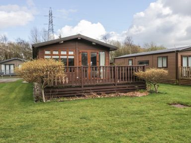 A wooden cabin with a front porch and railing surrounded by grass and small trees at Gressingham View Lodge Dock Acres near Carnforth