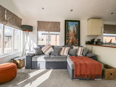 A living room with a gray sectional sofa with cushions and a red throw blanket a floor lamp an orange ottoman a side table and windows at Gressingham View Lodge in Dock Acres near Carnforth