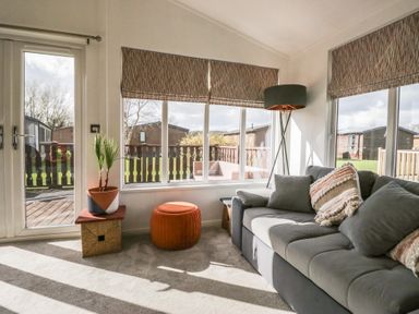 A living room with a gray sofa and patterned cushions near large windows with blinds a floor lamp a plant in a pot and an orange ottoman at Gressingham View Lodge Dock Acres near Carnforth