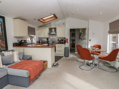 A living area with grey sofa and orange chairs around a glass table near a kitchen with cream cabinets at Gressingham View Lodge Dock Acres near Carnforth