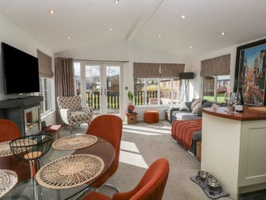 A living room with a glass dining table and orange chairs a gray sofa with a red throw a patterned armchair a flat screen tv a fireplace and large windows at Gressingham View Lodge in Dock Acres near Carnforth