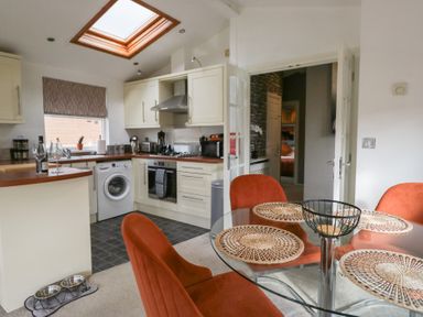 A kitchen with cream cabinets and appliances next to a dining area with a glass table and orange chairs at Gressingham View Lodge Dock Acres near Carnforth