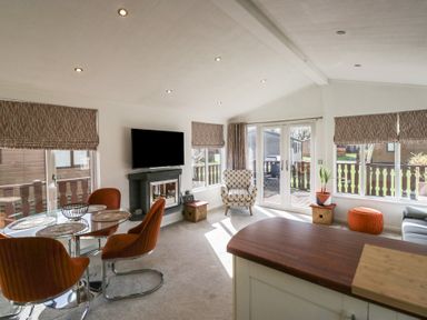 A living room with orange chairs around a glass table a patterned armchair a wall-mounted TV and patio doors at Gressingham View Lodge Dock Acres near Carnforth