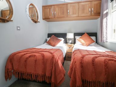 A bedroom with two single beds orange throws and pillows a bedside table lamp wooden cupboards and two round mirrors at Gressingham View Lodge in Dock Acres near Carnforth