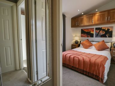 A bedroom with a bed with orange pillows and a blanket two bedside tables with lamps wooden cabinets above the bed and landscape pictures on the wall at Gressingham View Lodge in Dock Acres near Carnforth