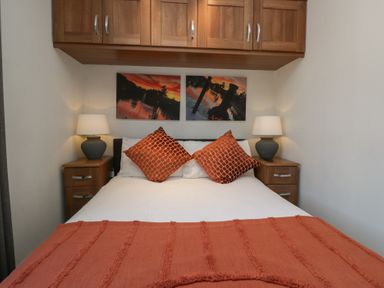 A bedroom with a bed with orange pillows and blanket two bedside tables with lamps and wooden cabinets above the bed at Gressingham View Lodge in Dock Acres near Carnforth