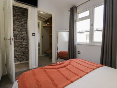 A bedroom with a bed covered with an orange blanket a mirror near the window and two doorways showing a walk-in wardrobe and a bathroom at Gressingham View Lodge in Dock Acres near Carnforth