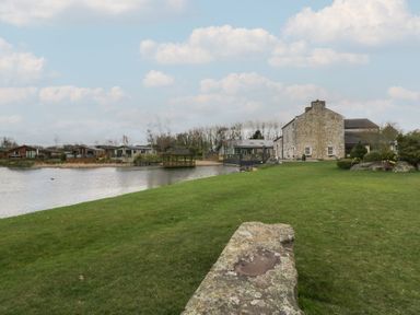 A stone house next to a lake with green lawn and wooden gazebos at Gressingham View Lodge in Dock Acres near Carnforth