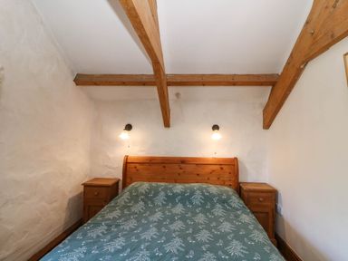 A bed with a wooden headboard and floral bedspread between two wooden nightstands with wall lamps above in a room with wooden ceiling beams at Cove Cottage in Llangrannog