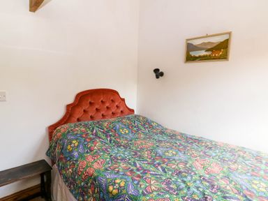 A bedroom with a bed covered by a butterfly patterned bedspread a red tufted headboard a wooden bench and a framed landscape painting on the wall at Cove Cottage in Llangrannog