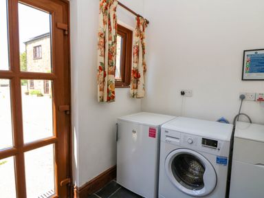 A laundry room with a washing machine and a small freezer next to a wooden door and a window with floral curtains at Cove Cottage in Llangrannog