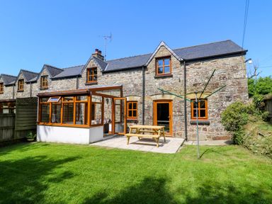 A stone house with wooden framed windows and doors a patio with a picnic table a glass conservatory and a grassy yard at Cove Cottage in Llangrannog