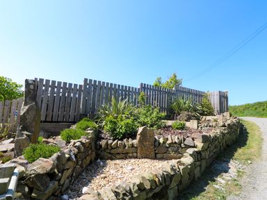 A garden with stone walls wooden fence and various plants next to a road at Cove Cottage in Llangrannog