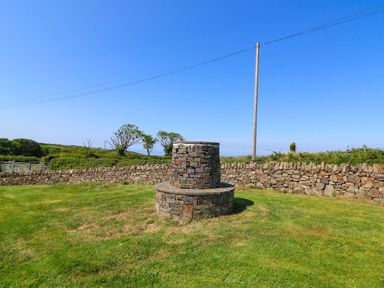 A stone structure on a grassy area with a stone wall and trees in the background at Cove Cottage in Llangrannog