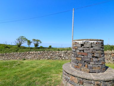 An outdoor scene with a stone well or structure on grass and a stone wall in the background at Cove Cottage in Llangrannog