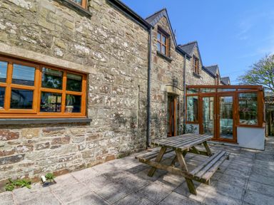 An outdoor patio area with a wooden picnic table next to a stone building with wooden windows and a glass conservatory at Y Bwthyn in Llangrannog