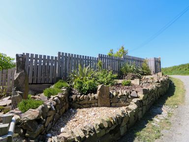 A stone garden planter with various plants and bushes behind a wooden fence along a gravel path at Y Bwthyn in Llangrannog