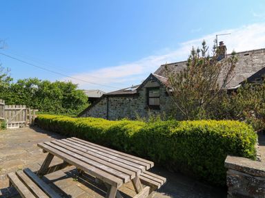 An outdoor patio area with a wooden picnic table and stone paving next to a stone house with a hedge and blue sky at Coach House in Llangrannog