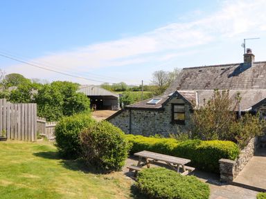 A stone house with a slate roof next to a garden with bushes and a wooden picnic table and a shed in the background at Coach House in Llangrannog