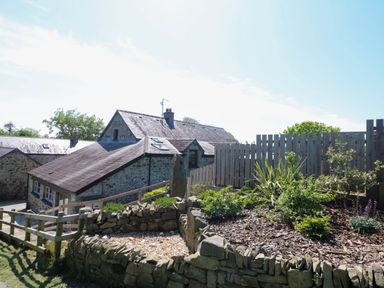 Stone buildings with slate roofs surrounded by a wooden fence and garden beds with plants at Coach House in Llangrannog