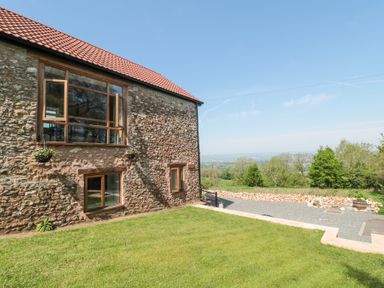 Stone cottage with wooden window frames and flower pots on a grassy lawn with trees and countryside in the background at Newlands Barn in Wellington