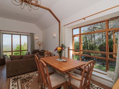A dining table with chairs and a vase of flowers next to large windows and a seating area with sofas in a room at Newlands Barn in Wellington